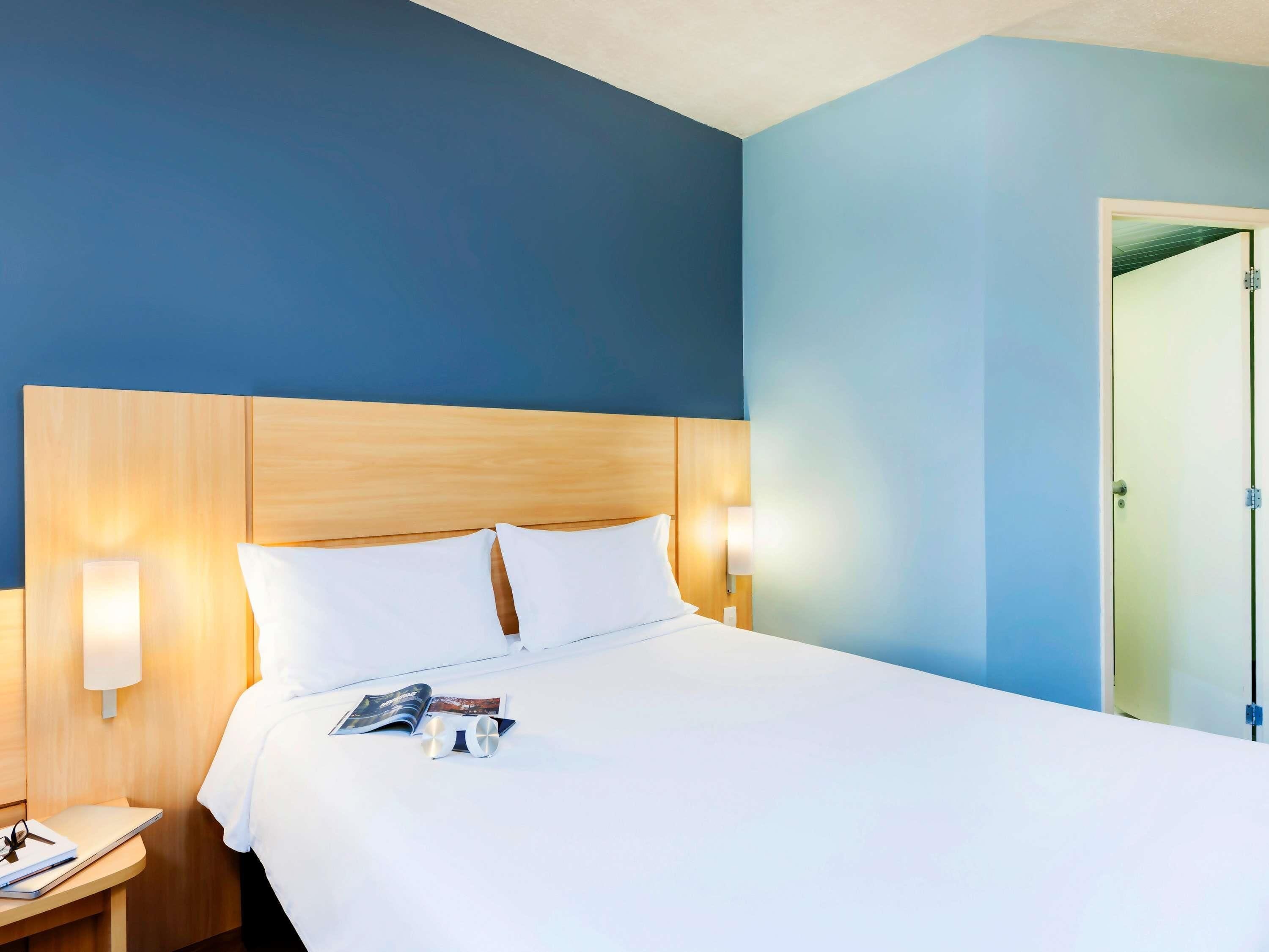 Ein Hotelzimmer mit Holzkopfteil, einem Bett mit weißer Bettwäsche, einem Nachttisch mit Lampe und einer Tür rechts.