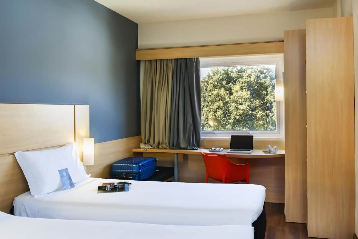 Ett hotellrum med en säng, ett skrivbord med en bärbar dator, en röd stol, en resväska och ett fönster med gardiner.
