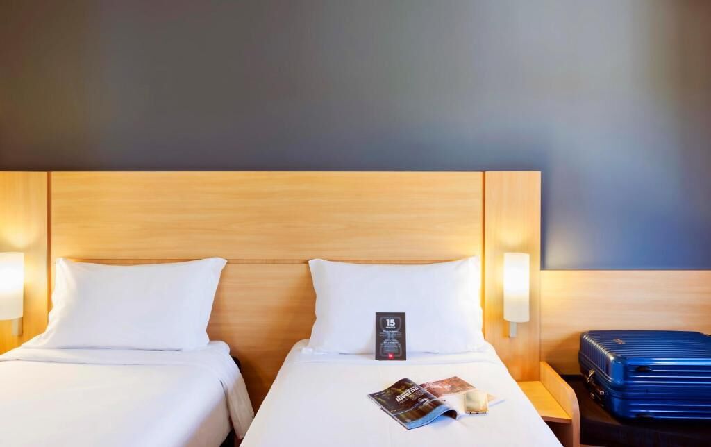 A hotel room with two beds, a wooden headboard, bedside lamps, a clock, and a blue suitcase on the right side.