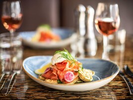 Un plato de comida con salmón ahumado, un huevo escalfado y adorno de eneldo, acompañado de una copa de vino rosado.