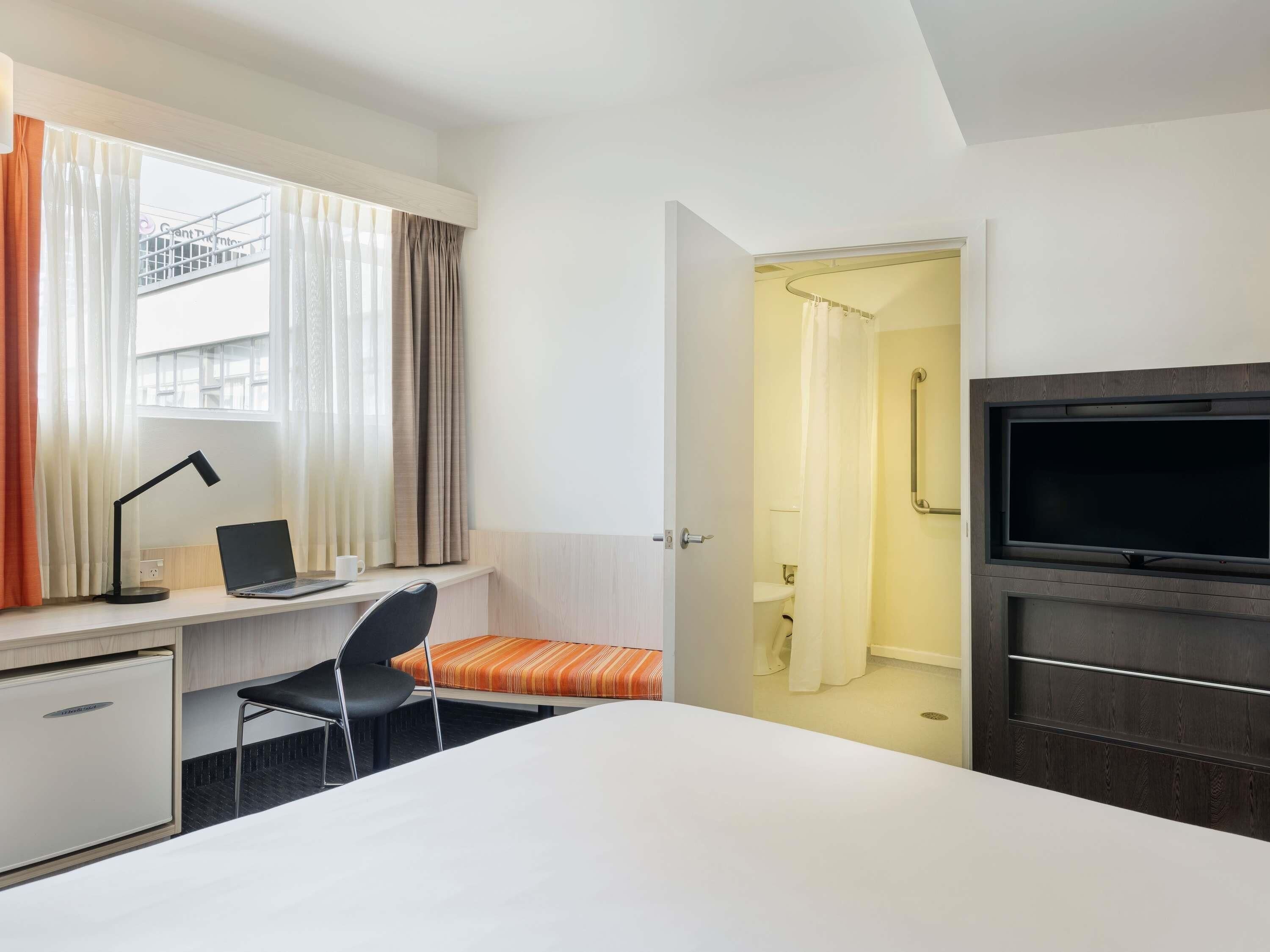 A hotel room with a bed, a desk with a laptop, a chair, a TV, and a shower area.