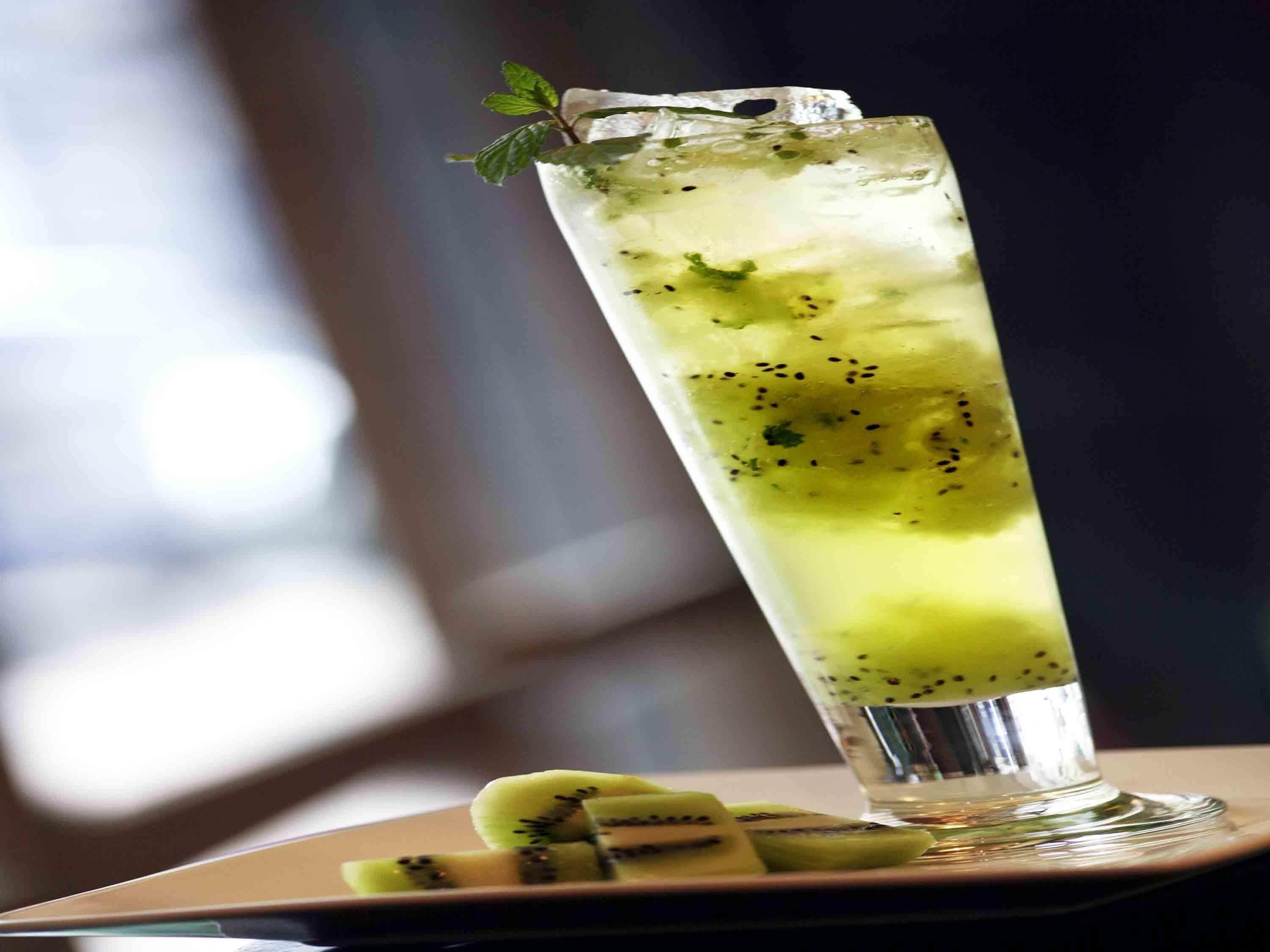 A tall glass with a green beverage, ice, and mint garnish, placed on a plate with kiwi slices.