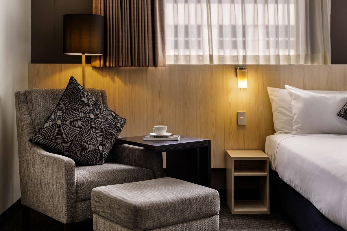 A hotel room with a bed, a chair with patterned cushions, a small table with a cup, and a nightstand.