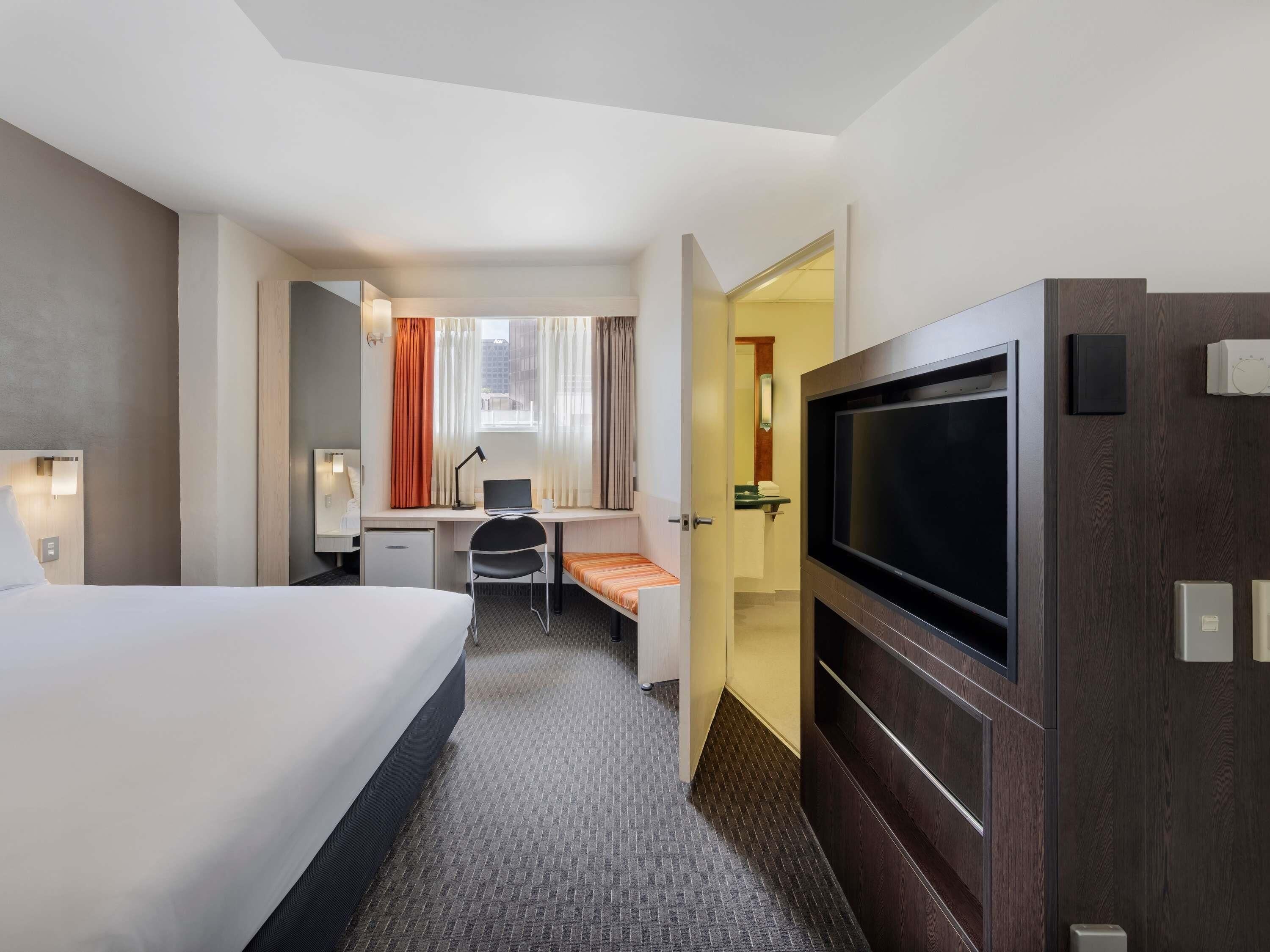 Habitación de hotel con cama, escritorio, silla, televisor y un baño visible a través de una puerta abierta.