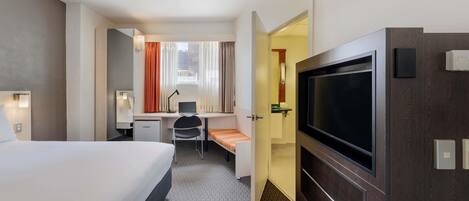 Habitación de hotel con cama, escritorio, silla, televisor y un baño visible a través de una puerta abierta.