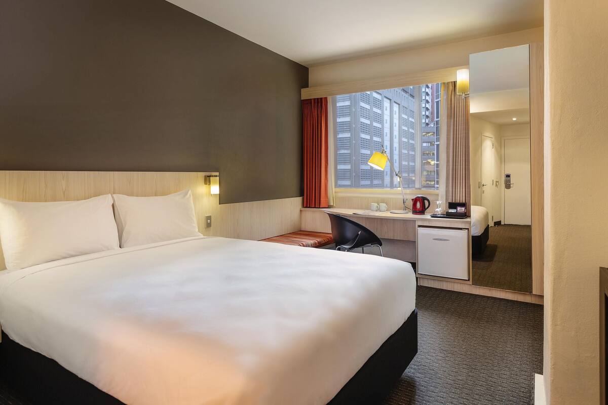 A hotel room with a large bed, a desk with a lamp, a coffee maker, and a view of tall buildings through the window.