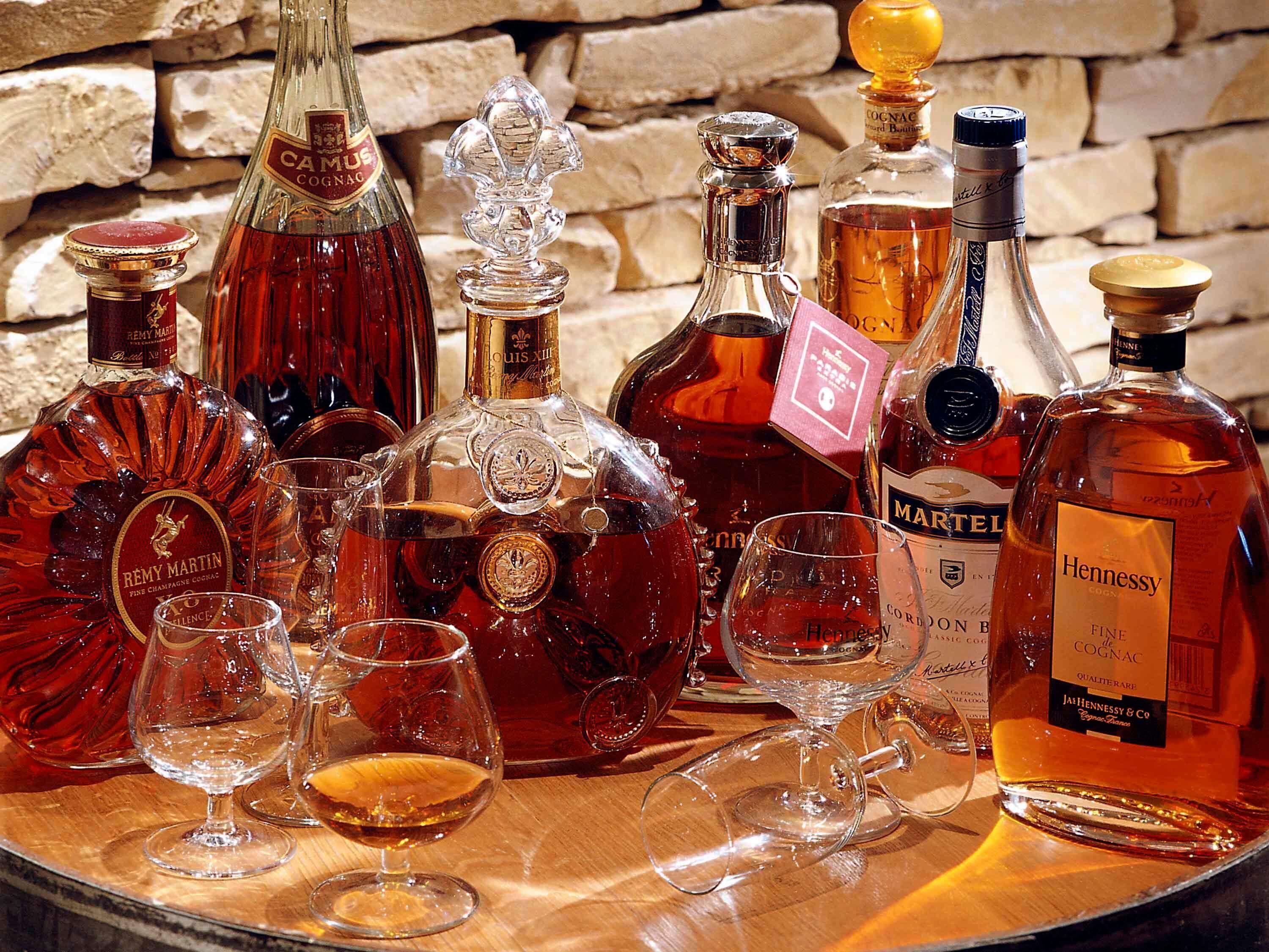 A collection of liquor bottles and glasses on a wooden table against a stone wall.