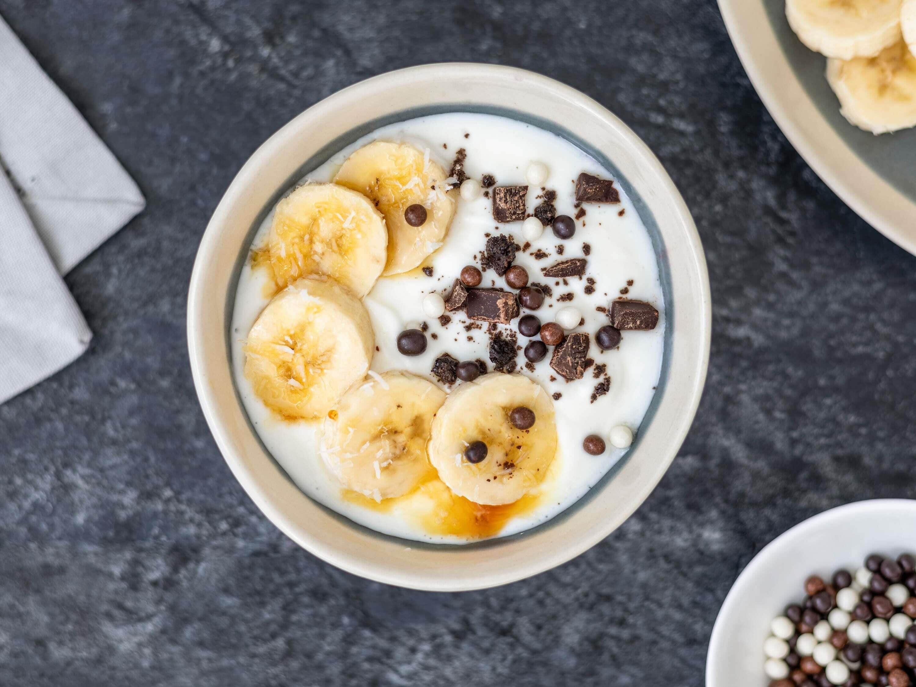 A bowl of yogurt with banana slices, chocolate pieces, and coconut flakes.
