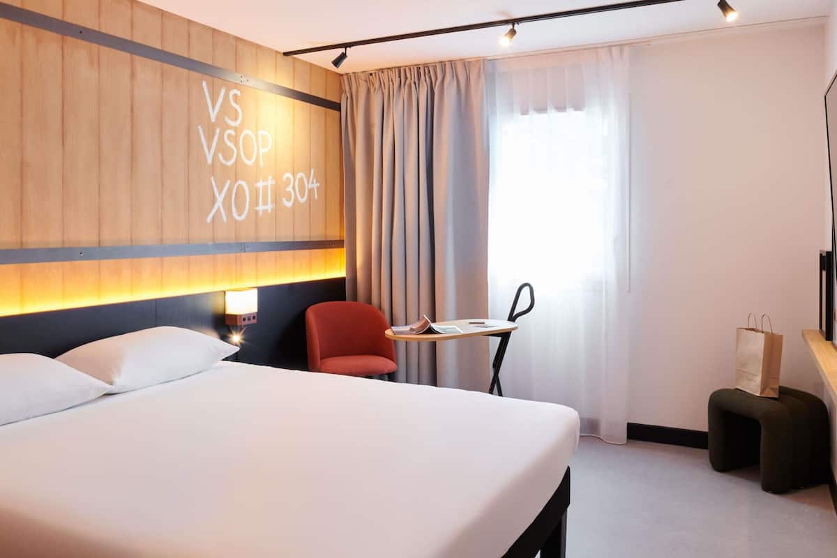 Ett hotellrum med en stor säng, ett skrivbord med en stol, ett litet bord och ett fönster med gardiner.