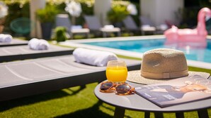 Ein Poolbereich mit Liegestühlen, einem Hut, Sonnenbrillen, einem Glas Orangensaft und einer Zeitschrift auf einem Tisch.