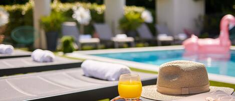 A poolside area with lounge chairs, a hat, sunglasses, a glass of orange juice, and a magazine on a table.