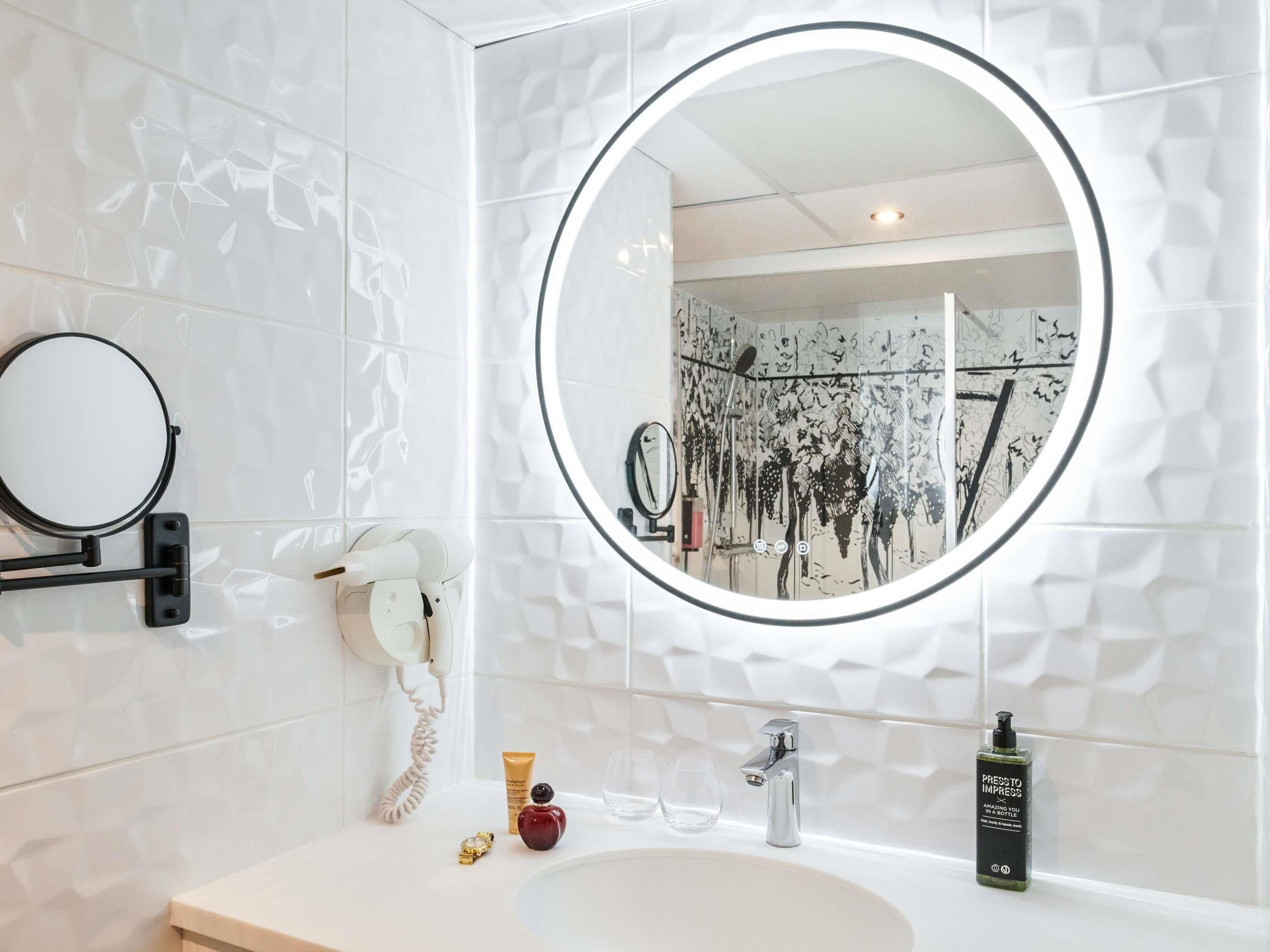 A bathroom with a round mirror, a white sink, a handheld showerhead, a telephone, and a bottle of shampoo.