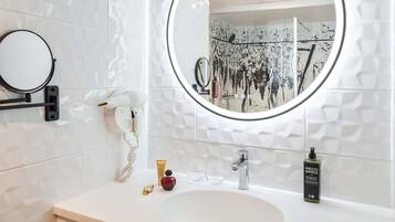 Een badkamer met een ronde spiegel, een witte wastafel, een handdouche, een telefoon en een fles shampoo.
