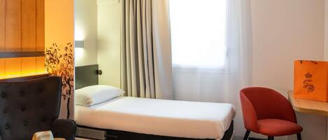 A hotel room with a bed, a chair, a small table, a wardrobe, and a window with curtains.