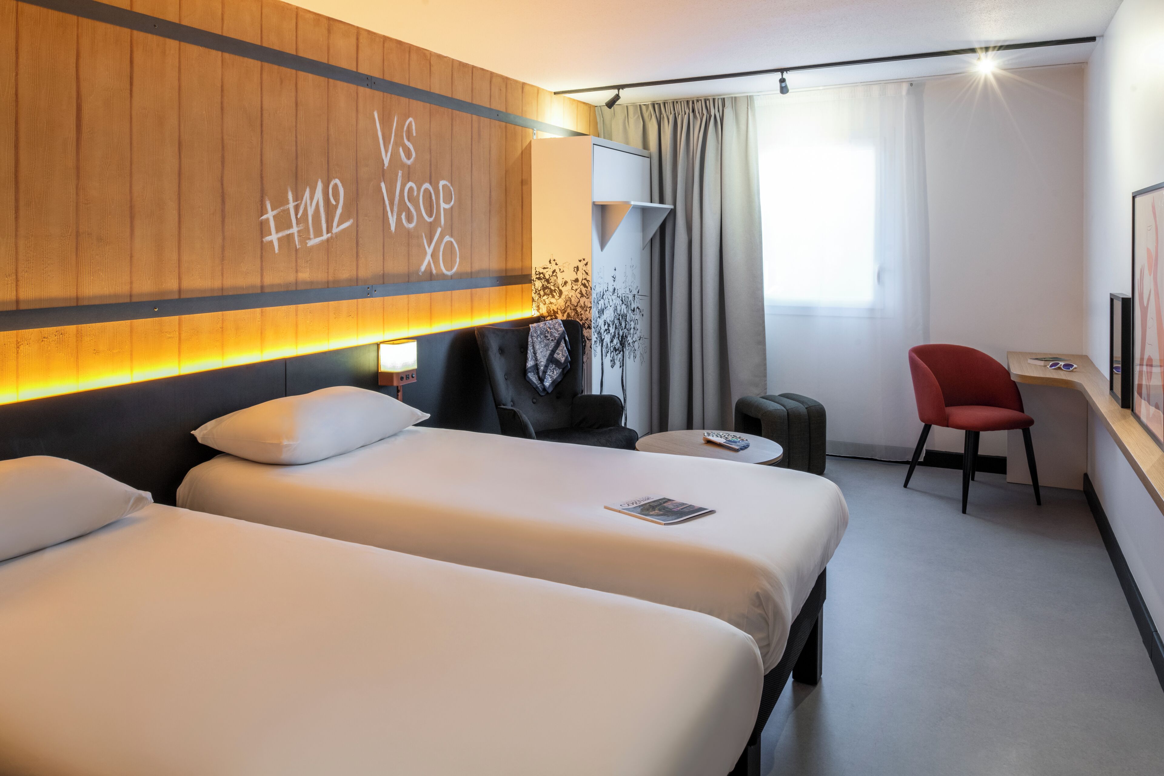 A hotel room with two beds, a desk, a red chair, and a wall with a wooden panel and text.