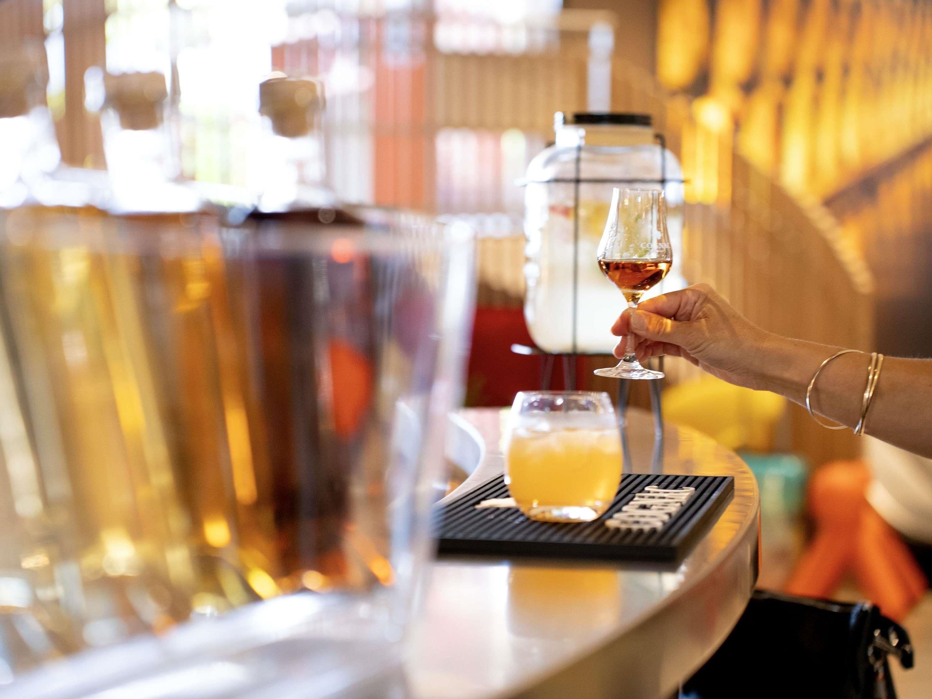 Zona de bar con una persona sosteniendo una copa de vino, una grifería y un vaso de jugo de naranja sobre una alfombrilla.