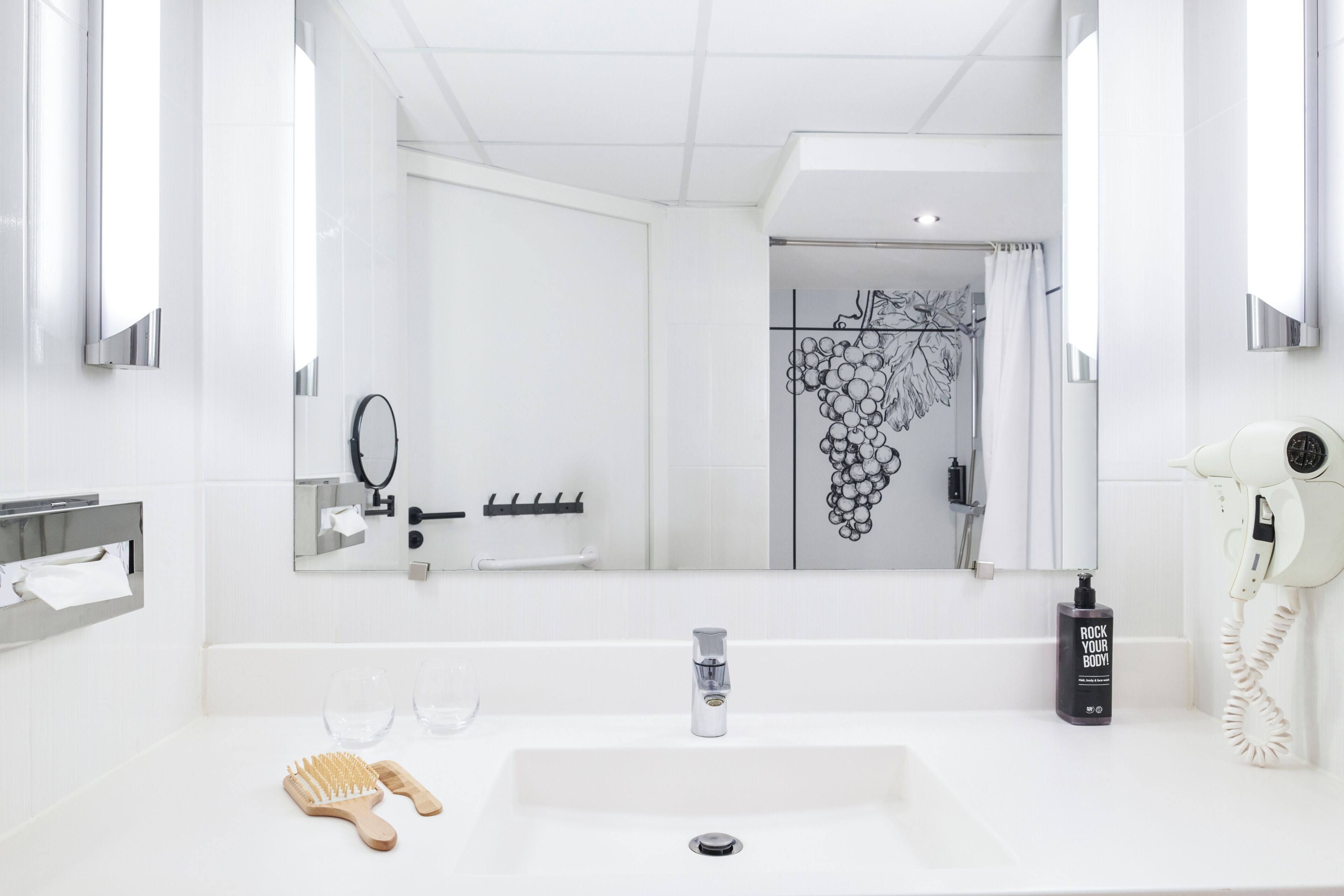 A modern bathroom with a large mirror, a sink, a hairdryer, and a bottle of shampoo.