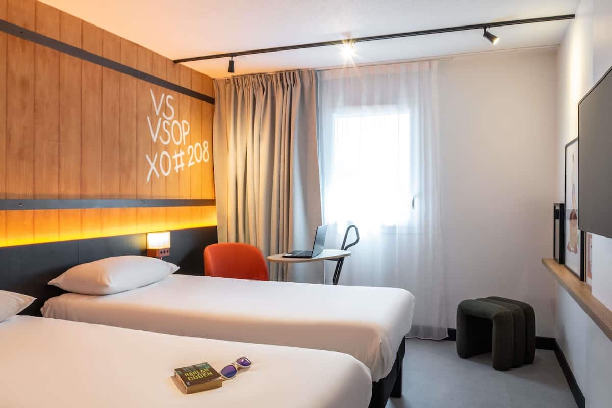Ett hotellrum med två sängar, ett skrivbord med en bärbar dator, en stol, en tv och ett fönster med gardiner.