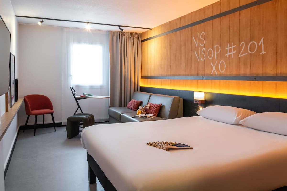 Ett hotellrum med en säng, ett skrivbord, en stol, en resväska och en vägg med en skylt.
