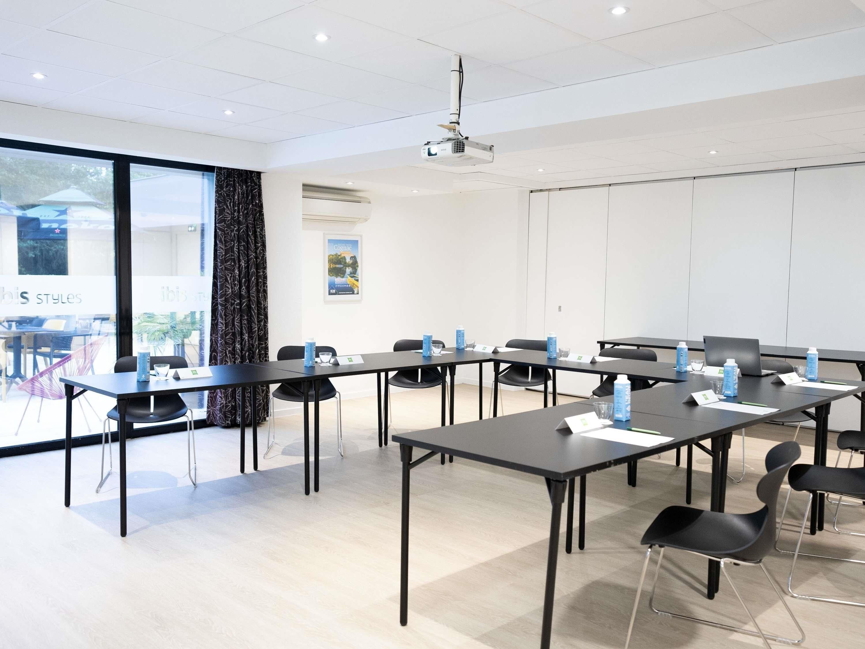 Une salle de conférence équipée de tables, de chaises, d’un projecteur et d’une grande fenêtre.