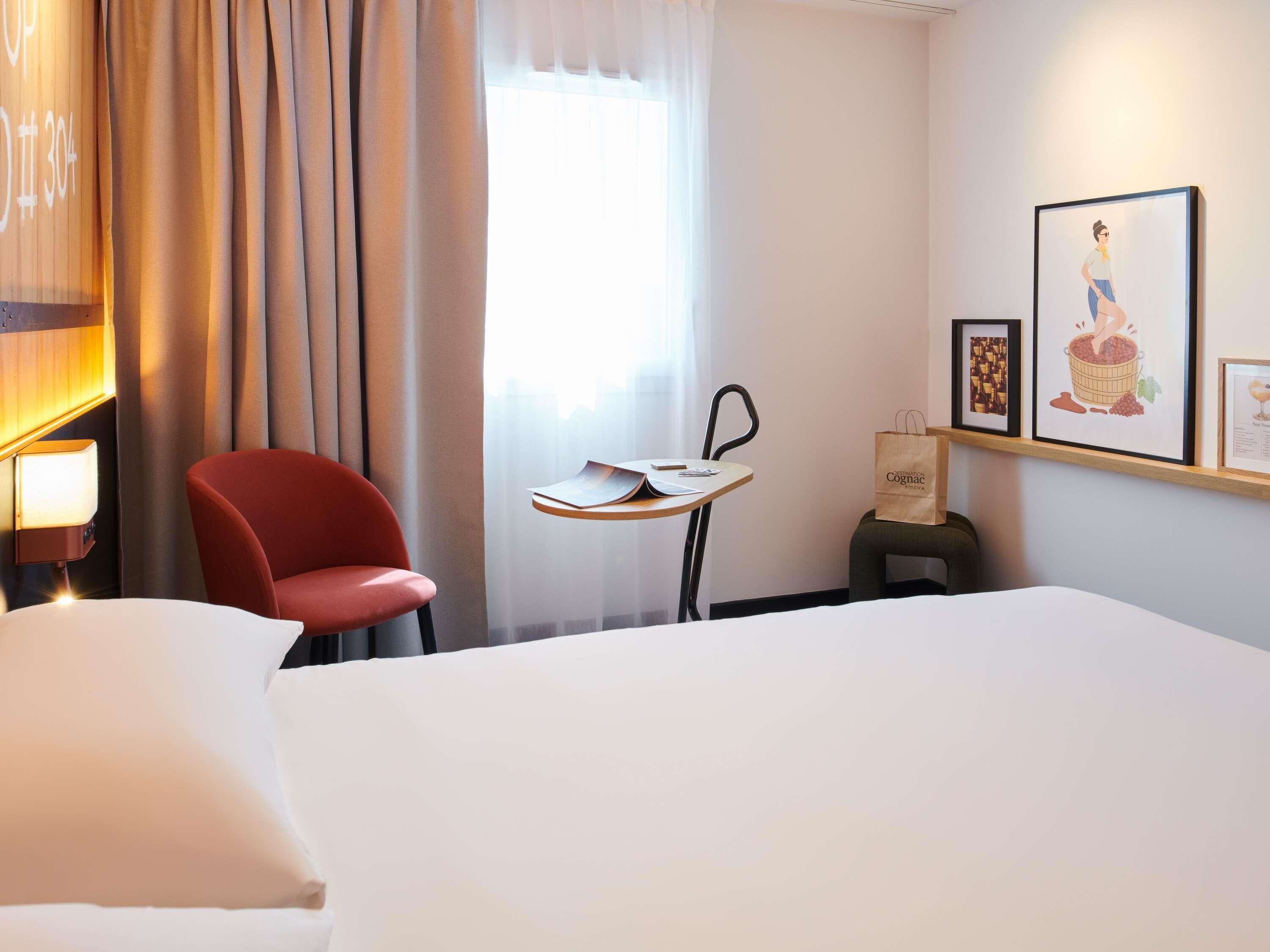 A hotel room with a bed, a red chair, a small table with a lamp, and a window with curtains.