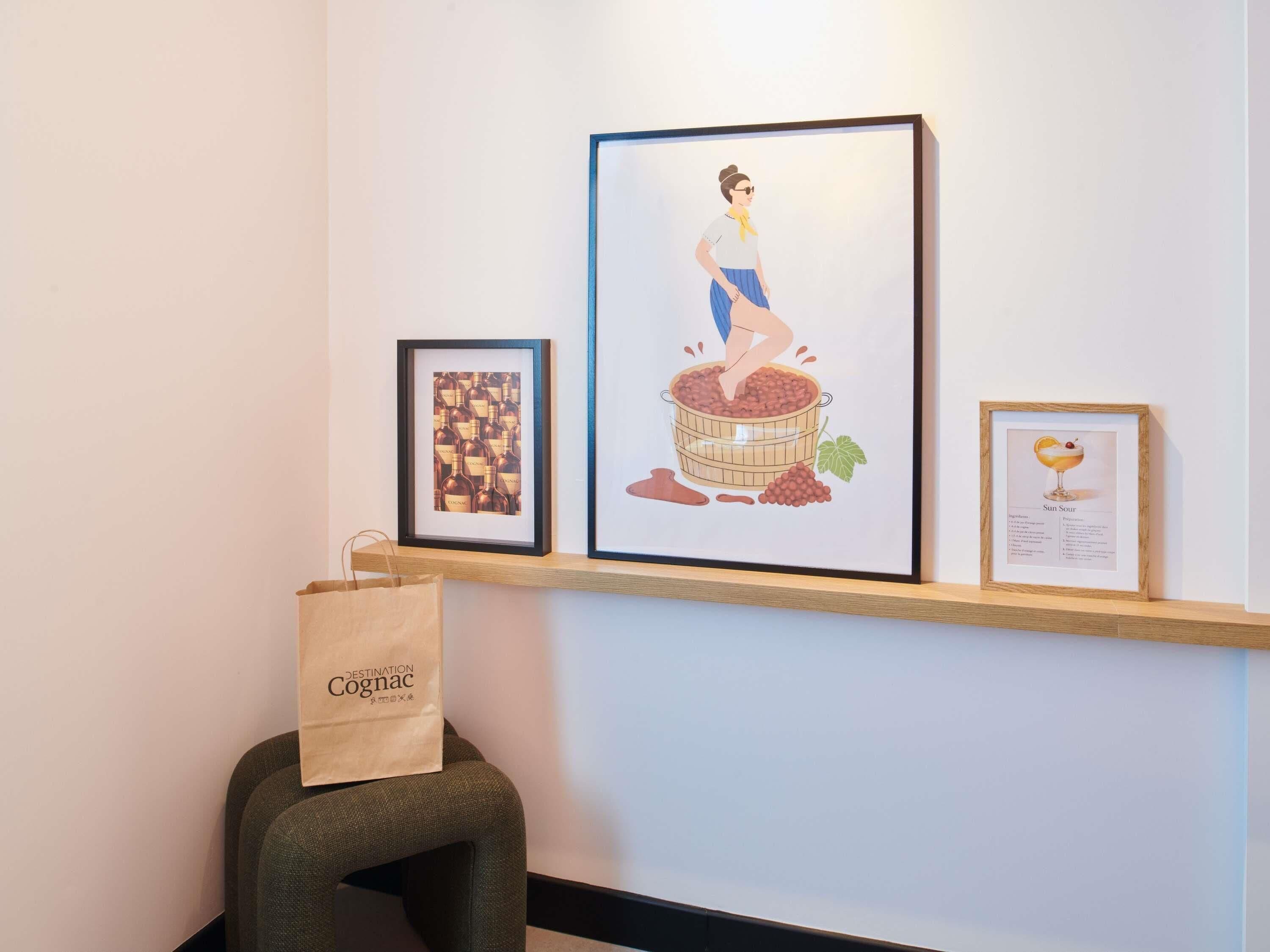 A wall with three framed artworks, a wooden shelf, and a paper bag on a stool.