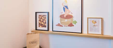 A wall with three framed artworks, a wooden shelf, and a paper bag on a stool.