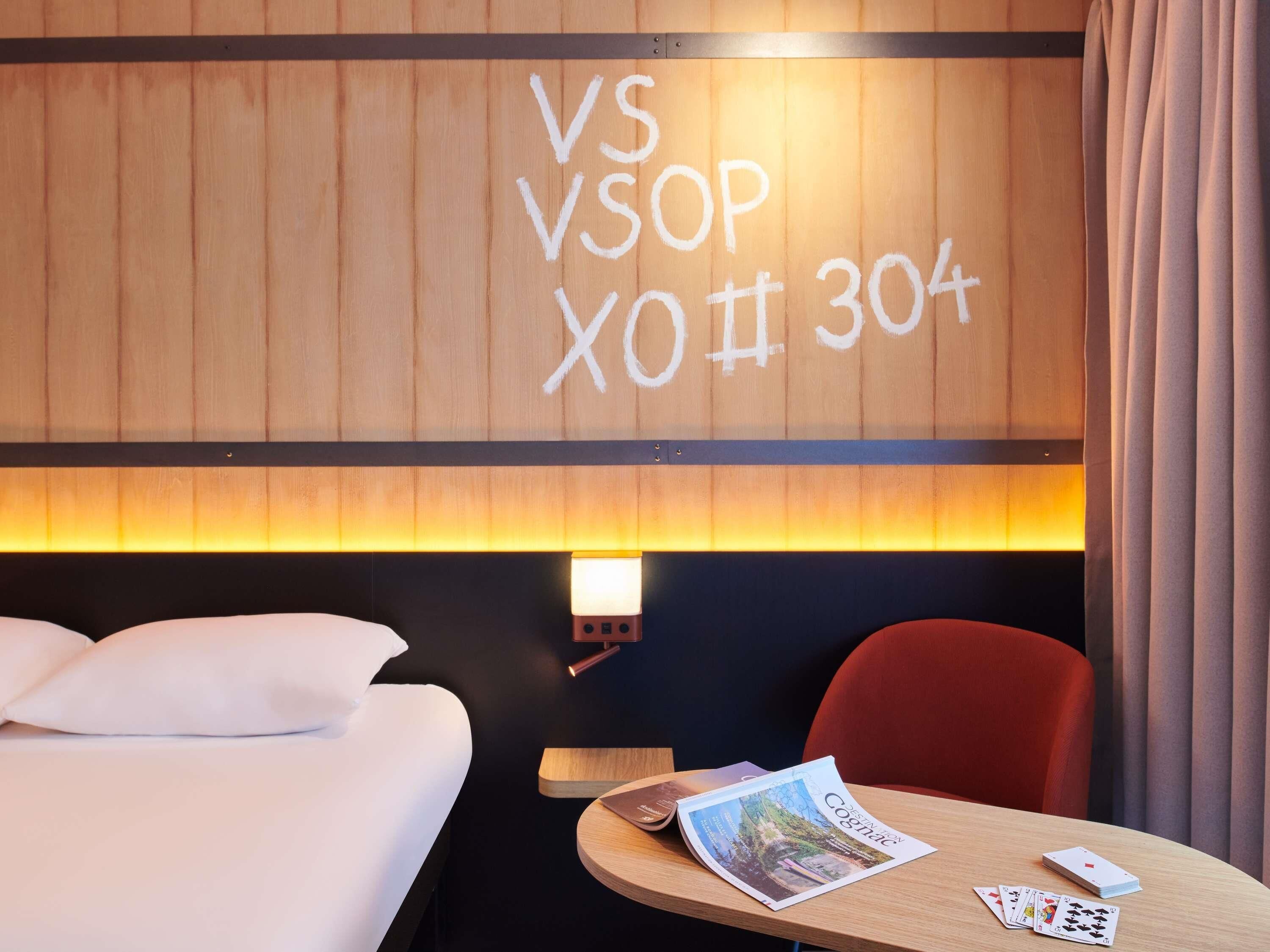 A hotel room with a bed, a table with a magazine and playing cards, and a wall with 'VS VSOP XO 304+' written on it.