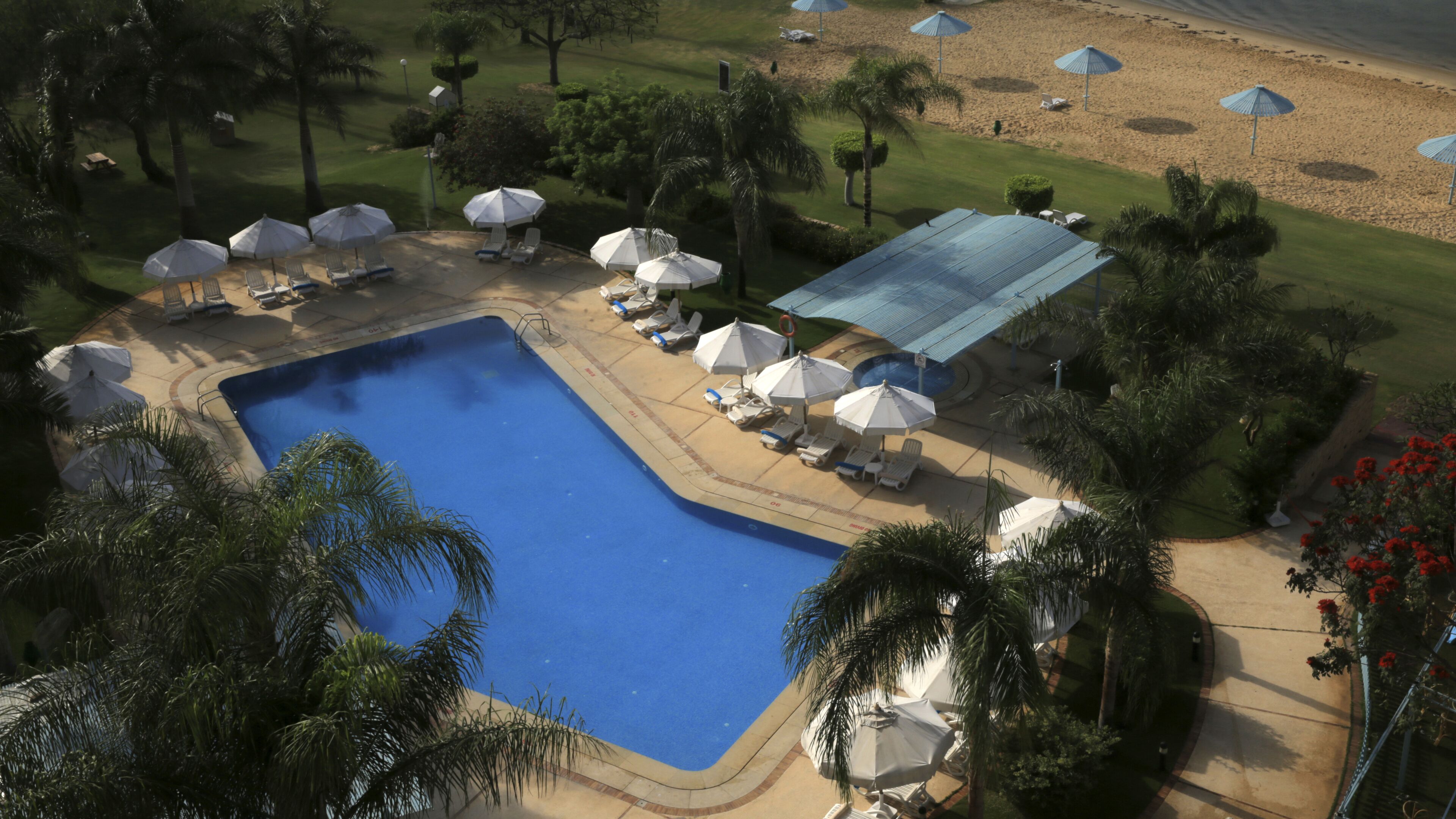 An aerial view of a resort with a central swimming pool, surrounded by lounge chairs and umbrellas, with a golf course in the background.