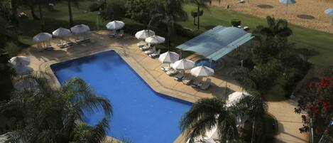 An aerial view of a resort with a central swimming pool, surrounded by lounge chairs and umbrellas, with a golf course in the background.