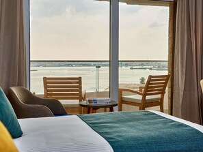 A hotel room with a large window, two wooden chairs, a small table, and a bed with a blue blanket. - Mercure Ismailia Forsan Island Hotel (Ismailia)
