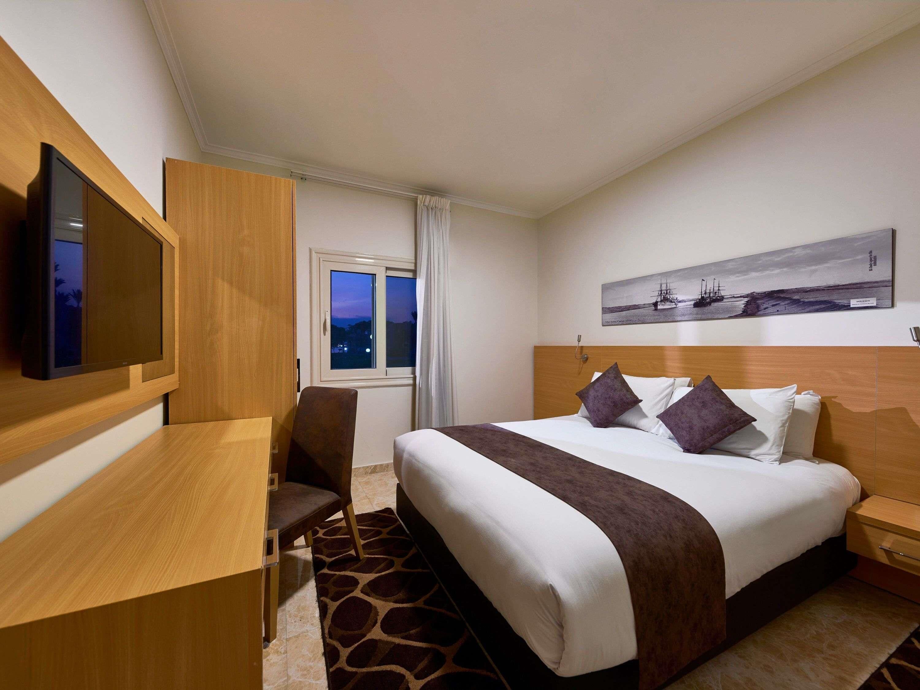 Quarto de hotel com cama, escrivaninha, cadeira, televisão, janela e um quadro na parede.