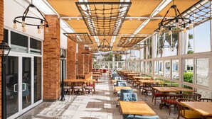 Un café al aire libre con paredes de ladrillo, mesas y sillas de madera, suelo de baldosas con diseño y techo de red de cuerda.