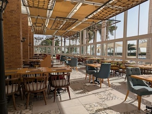 A spacious indoor dining area with multiple tables and chairs, large windows offering views of palm trees and the sea, and a brick wall on one side.