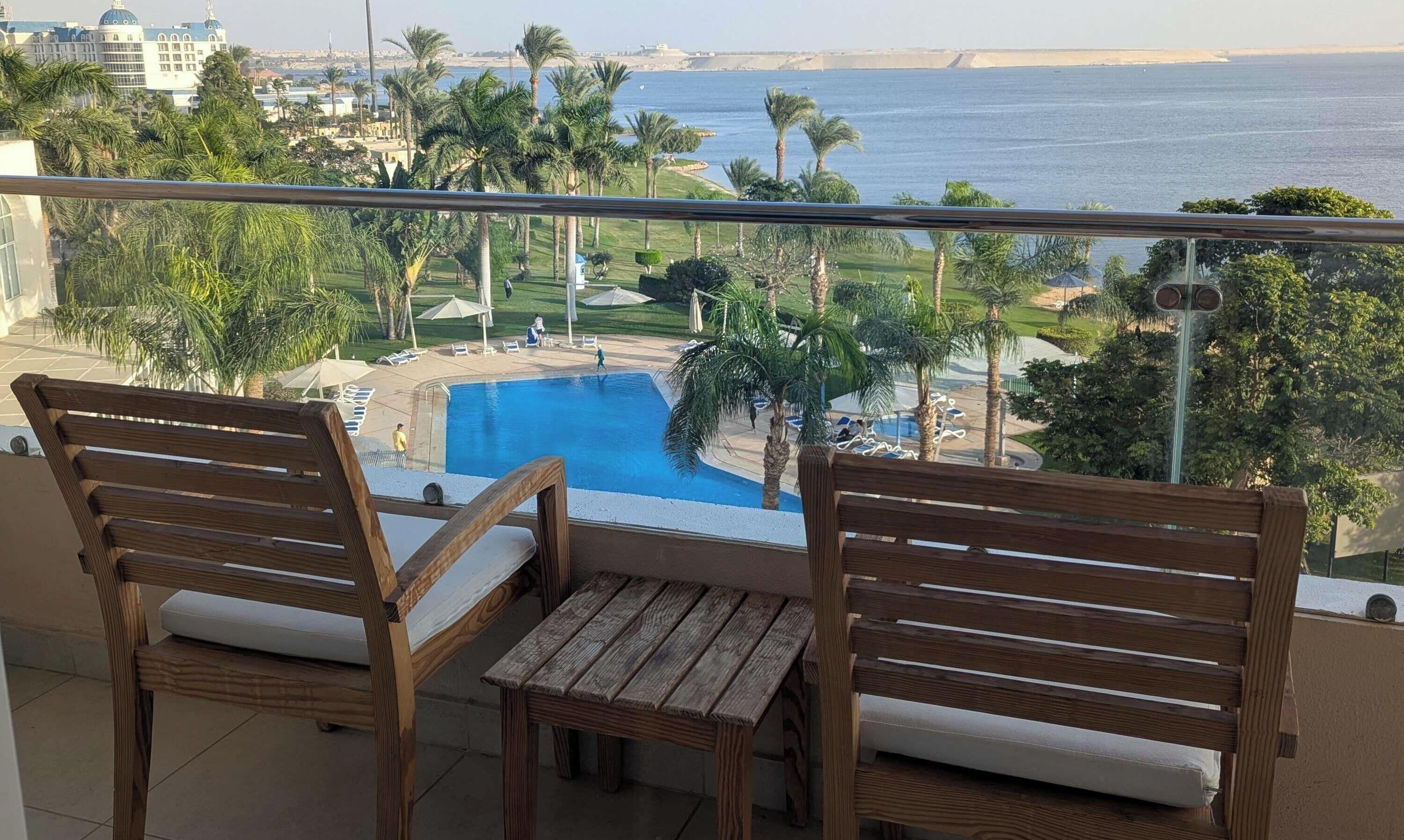 A balcony with wooden chairs and a small table overlooking a swimming pool and a coastal view.