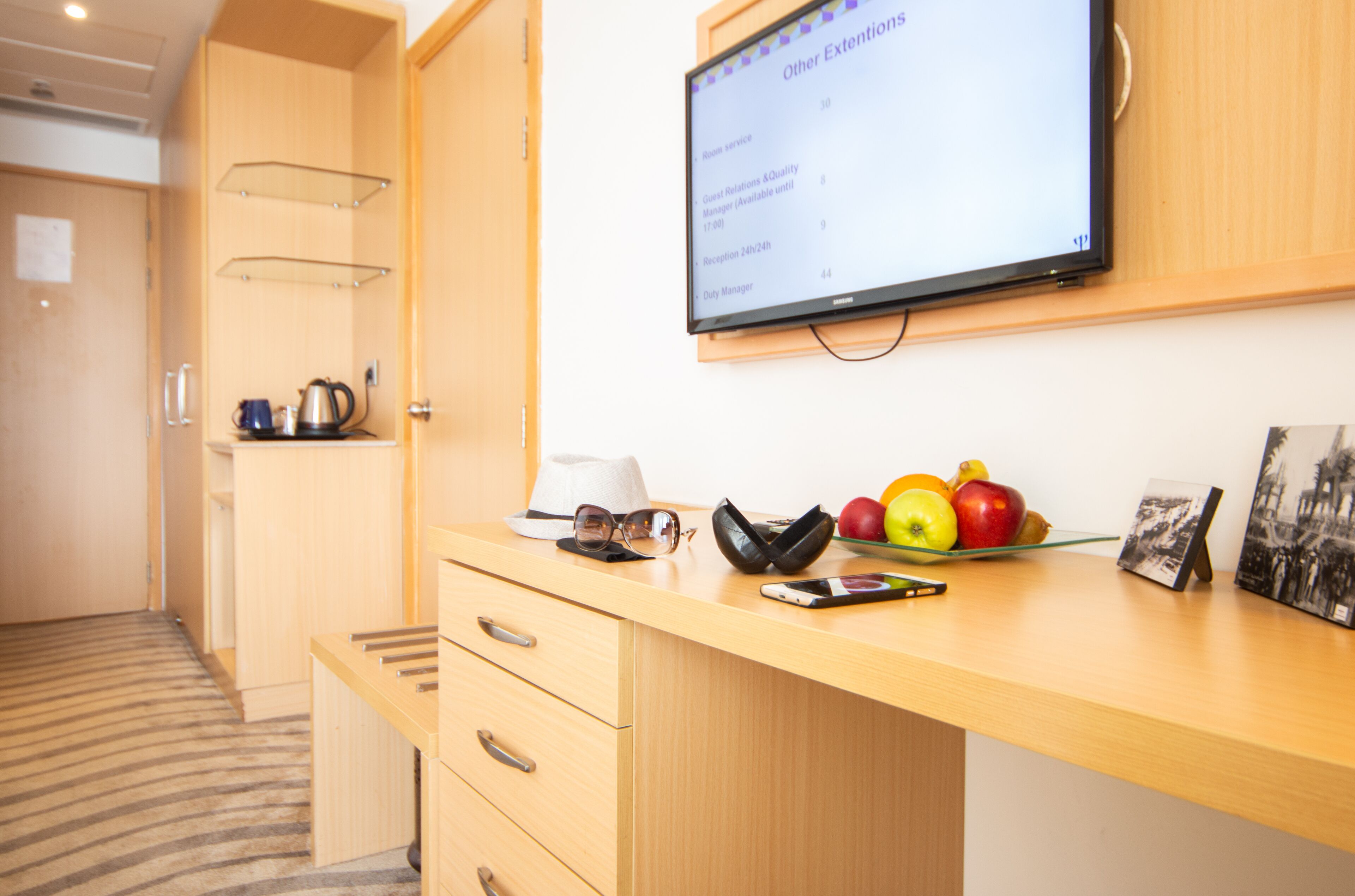 Mostrador de recepción de hotel con un monitor mostrando opciones de servicio a la habitación, un frutero, un sombrero, gafas de sol, una billetera y una foto enmarco.
