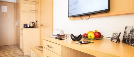 Mostrador de recepción de hotel con un monitor mostrando opciones de servicio a la habitación, un frutero, un sombrero, gafas de sol, una billetera y una foto enmarco.