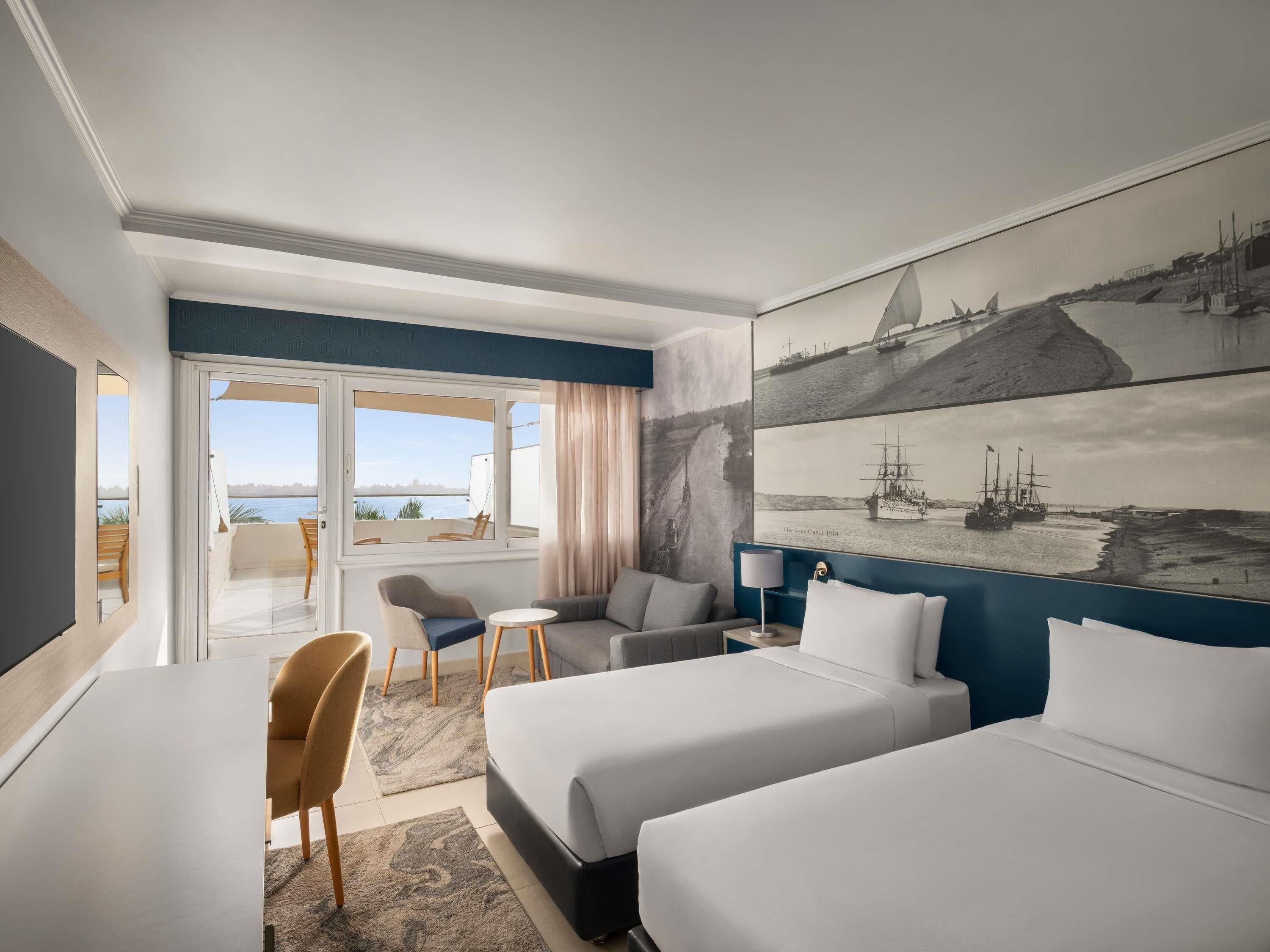 Quarto de hotel com duas camas, uma escrivaninha, uma cadeira, uma mesinha e uma janela grande com vista para o mar.