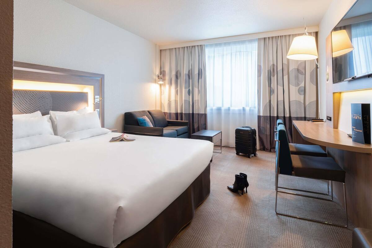 A hotel room with a large bed, a desk, a chair, a sofa, a suitcase, and a television.