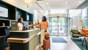 Un hall d’hôtel à l’aménagement moderne, avec un comptoir d’accueil, une porte tournante et un espace salon offrant une vue sur l’extérieur.