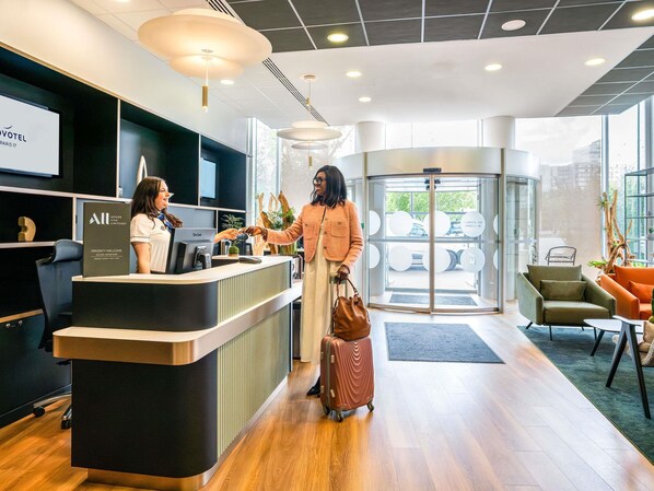 Zona de recepción de hotel con diseño moderno, una mesa de recepción, un giratorio y un área de descanso con vistas al exterior.