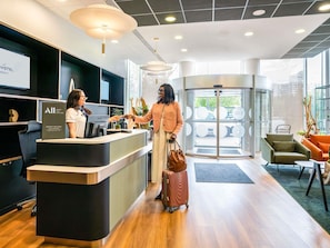 A hotel reception area with a modern design, a reception desk, a revolving door, and a seating area with a view of the outside. - Novotel Paris 17 (Paris)