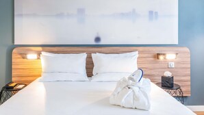 Une chambre d’hôtel avec un grand lit, deux oreillers, une tête de lit dotée d’un éclairage intégré, une œuvre d’art accrochée au mur et une table de chevet avec un téléphone et une boîte de mouchoirs.