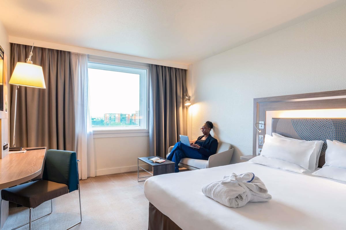 A hotel room with a bed, a desk, a chair, a lamp, and a view of buildings outside the window.