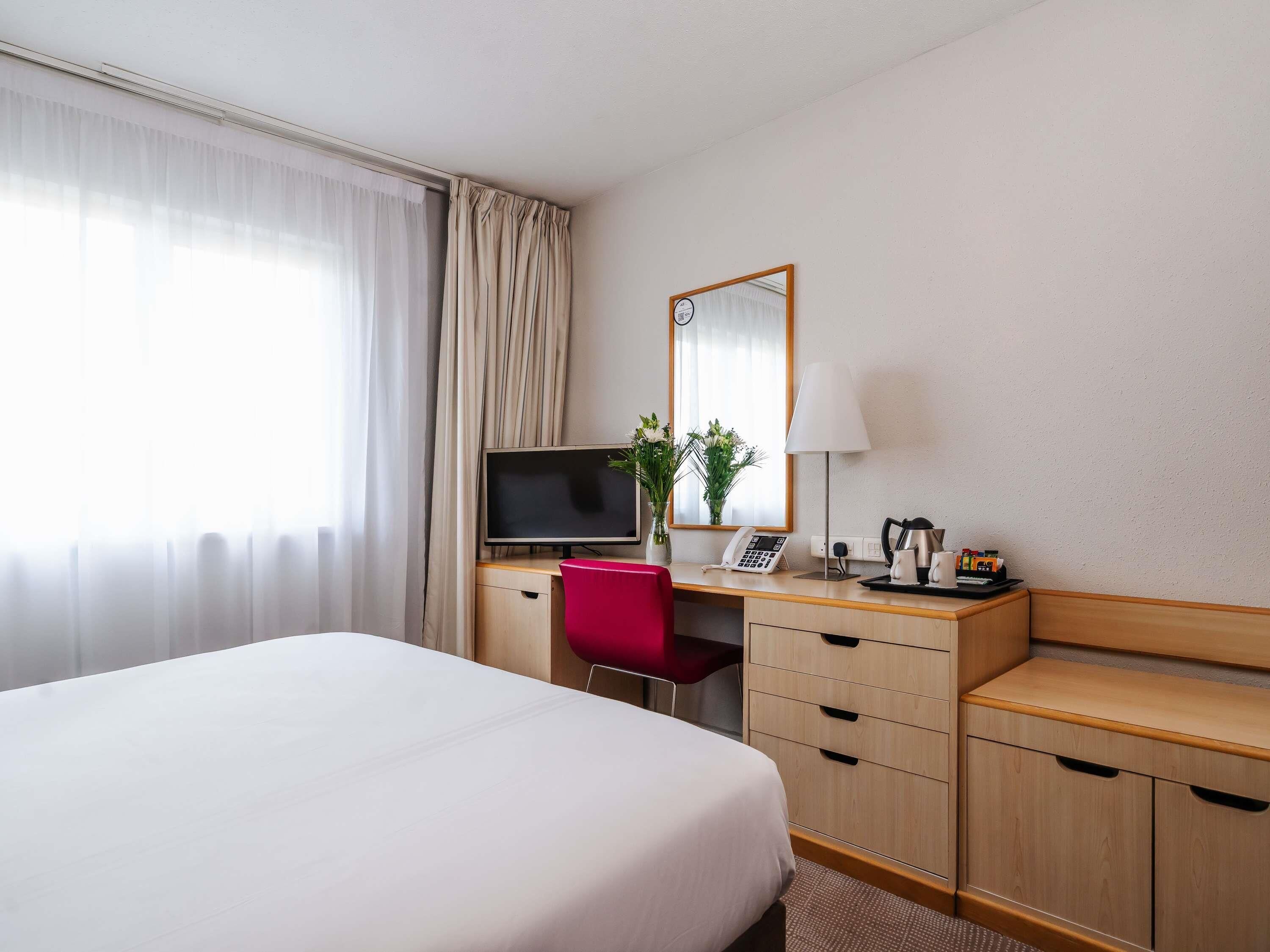 Et hotelværelse med seng, skrivebord med fjernsyn, stol, lampe, telefon og kaffemaskine.