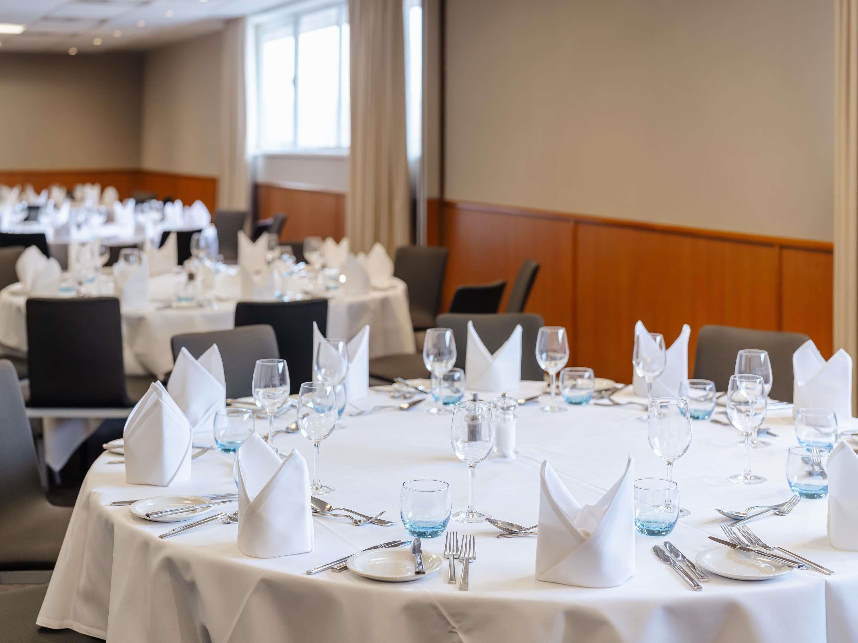 Un comedor de hotel con mesas redondas dispuestas para el servicio, cada una con mantel blanco, cubiertos y vasos de agua azules.