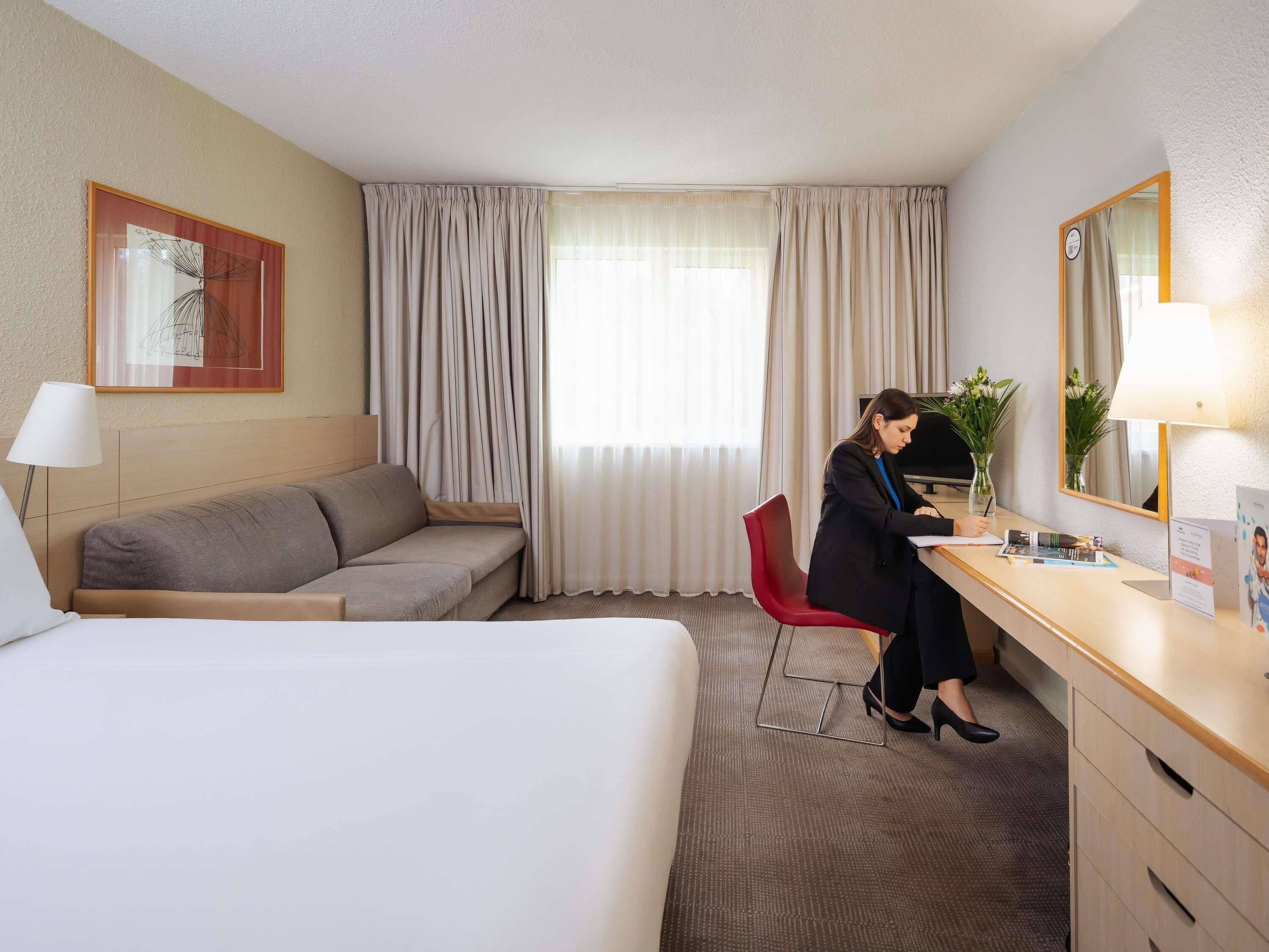 Ein Hotelzimmer mit einem Bett, einem Schreibtisch, einem roten Sessel, einer Couch, einem großen Fenster mit Vorhängen, einer Wanduhr und einem gerahmten Bild.