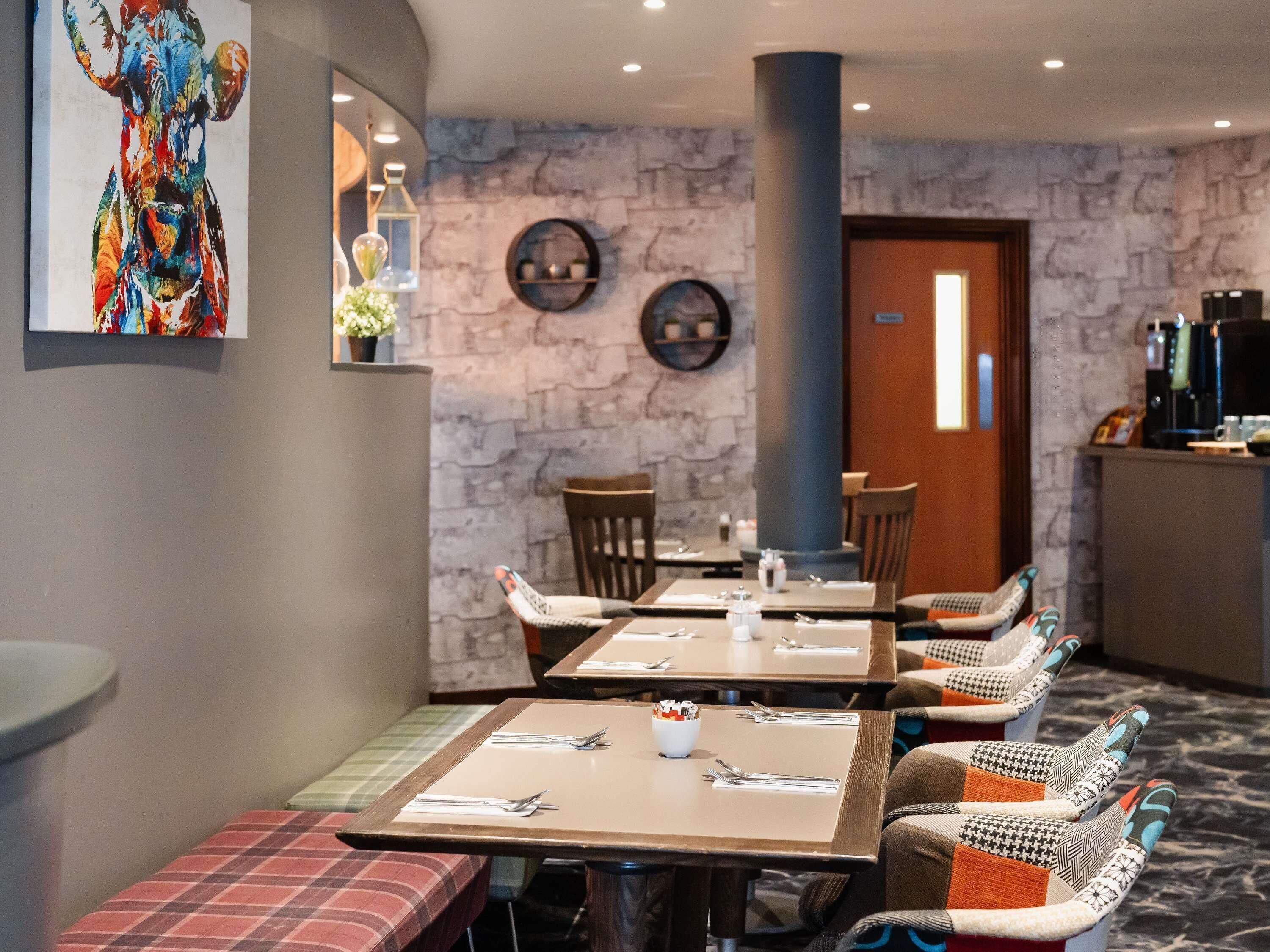 A dining area with tables set for four, benches, and a stone wall with a painting.