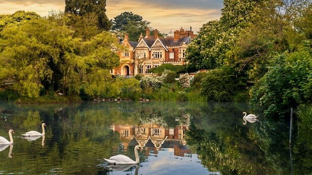 A large, historic building with a reflection in the water, surrounded by lush greenery and swans. - Novotel Milton Keynes (Milton Keynes)