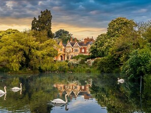 A large, historic building with a reflection in the water, surrounded by lush greenery and swans. - Novotel Milton Keynes (Milton Keynes)